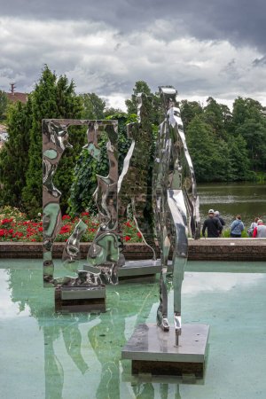 Foto de Finlandia, Kotka - 18 de julio de 2022: Parque Sopokanlahti y lago. Estatuas de arte de corte moderno de metal brillante en la piscina de agua verde. Cinturón verde bajo el paisaje nublado gris. Flores rojas añadir color - Imagen libre de derechos