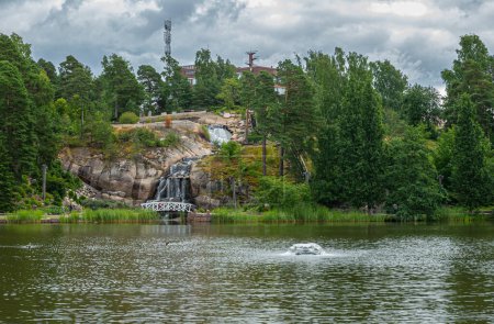 Photo for Finland, Kotka - July 18, 2022: Sopokanlahti park and lake. the waterfall from across the lake set in green environment under gray cloudscape. Building and antenna on horizon - Royalty Free Image