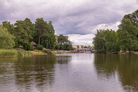 Photo for Finland, Kotka - July 18, 2022: Sopokanlahti park and lake. Whtie wooden bridge spans water link to harbor and sea under gray cloudscape. Green tree belt on both sides - Royalty Free Image