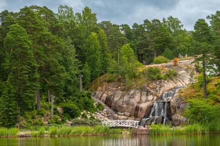 Photo for Finland, Kotka - July 18, 2022: Sopokanlahti park and lake. Side view on waterfall over rocks set in green foliage environment with small white wooden pedestrian bridge. Gray cloudscape - Royalty Free Image