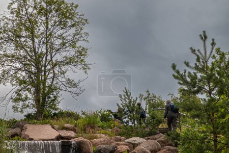 Photo for Finland, Kotka - July 18, 2022: Sopokanlahti park and lake. Family climbs stairs to top of waterfall between green foliage under dark cloudscape - Royalty Free Image