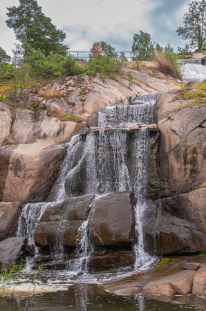 Photo for Finland, Kotka - July 18, 2022: Sopokanlahti park and lake. Closeup of lower half of waterfall producing white water over brown wet rocks. Some green foliage under blue cloudscape - Royalty Free Image