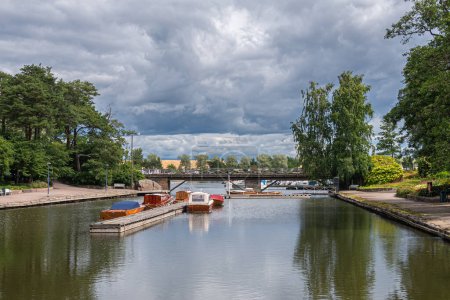 Photo for Finland, Kotka - July 18, 2022: Sopokanlahti park and lake. Access to harbor on waterway under heavy blueish cloudscape with green foliage on sides of Meriniementie bridge. Boats add colors - Royalty Free Image