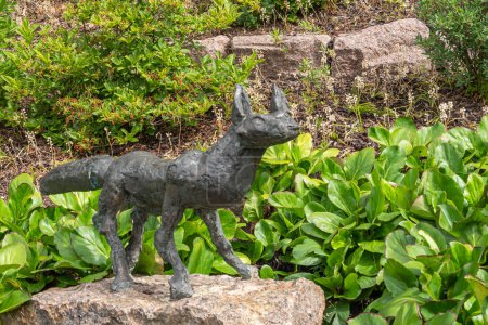 Photo for Finland, Kotka - July 18, 2022: Sopokanlahti park and lake. Closeup of bronze fox statue set on beige rock, surrounded by geen plant foliage. Tiny red flowers add color - Royalty Free Image