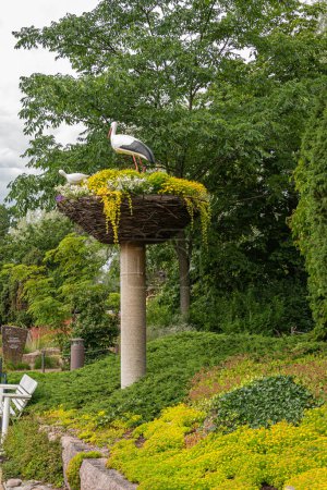 Photo for Finland, Kotka - July 18, 2022: Sopokanlahti park and lake. 2 Stork statues on top of fake nest now filled with abundance of yellow flowers. Set on column under green foliage. - Royalty Free Image