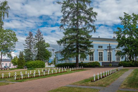 Photo for Finland, Hamina - July 18, 2022: WWII war cemetery with rows of white crosses and red flowers on lawn adjacent to blue St. Johns church, Johanneksen kirkko, under blue cloudscape - Royalty Free Image