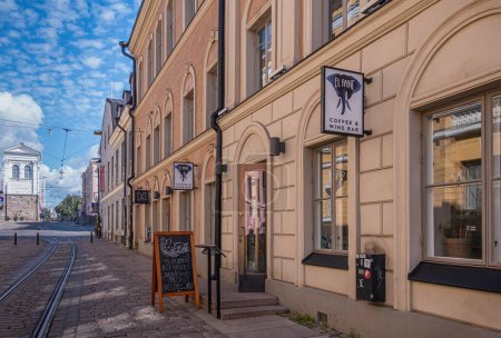Foto de Helsinki, Finlandia - 19 de julio de 2022: Fachada amarilla y entrada al bar El Fant Coffee and Wine en la calle Katariinankatu con rieles y bajo un paisaje nublado azul - Imagen libre de derechos
