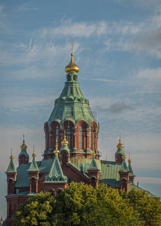 Foto de Helsinki, Finlandia - 19 de julio de 2022: Ladrillo rojo y techo verde La catedral ortodoxa rusa de Uspenski se eleva sobre árboles verdes bajo el cielo azul. Top dorado en cada torre - Imagen libre de derechos