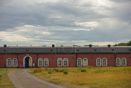 Foto de Helsinki, Finlandia - 19 de julio de 2022: Fortaleza Suomenlinna. Edificio histórico de larga residencia de piedra roja para soldados, hoy Centro Cultural Nórdico, HIAP, bajo un paisaje nublado gris, ubicado en un campo de pastos amarillos - Imagen libre de derechos