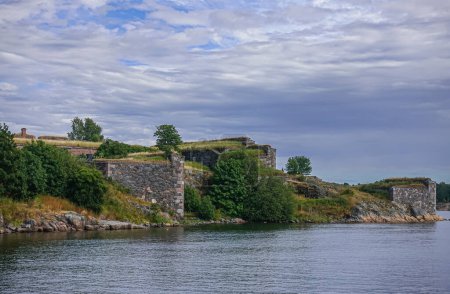 Foto de Helsinki, Finlandia - 19 de julio de 2022: Fortaleza Suomenlinna. Wrede Bastión murallas bajo azul-gris paisaje nuboso con vegetación verde cerca de la línea de agua. - Imagen libre de derechos
