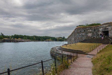 Foto de Helsinki, Finlandia - 19 de julio de 2022: Fortaleza Suomenlinna. escalones conducen a la puerta real a través de muralla de piedra gris desde el muelle de mar bajo el paisaje nublado gris. Follaje verde en el horizonte. Ropa añadir colores - Imagen libre de derechos
