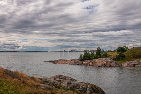 Foto de Helsinki, Finlandia - 19 de julio de 2022: Fortaleza Suomenlinna. Amplio paisaje en la ensenada del mar en la costa suroeste con Industria y paisaje urbano portuario de Munkkisaari bajo un paisaje nublado gris. Follaje verde por delante - Imagen libre de derechos