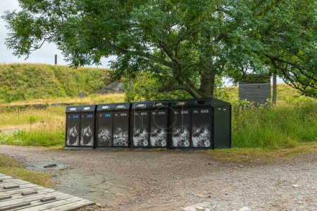 Foto de Helsinki, Finlandia - 19 de julio de 2022: Fortaleza Suomenlinna. Fila de basureros negros colocados a lo largo del camino de grava bajo un árbol de follaje verde - Imagen libre de derechos