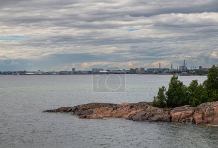 Foto de Helsinki, Finlandia - 19 de julio de 2022: Fortaleza Suomenlinna. Munkkisaarie puerto y distrito de la industria visto desde la costa oeste bajo nubes grises. Follaje verde en la costa de entrada de mar - Imagen libre de derechos