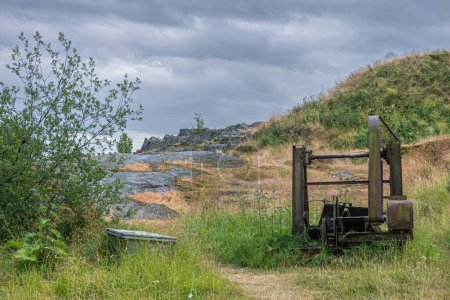 Foto de Helsinki, Finlandia - 19 de julio de 2022: Fortaleza Suomenlinna. Máquina de sierra pesada antigua dejada atrás cerca de la costa del mar en un entorno de malas hierbas verde bajo un paisaje nublado azul - Imagen libre de derechos