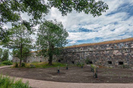 Foto de Helsinki, Finlandia - 19 de julio de 2022: Fortaleza Suomenlinna. Murallas de piedra gris cerca de Pipers Park bajo el paisaje nublado azul. Árboles nuevos plantados y otro follaje verde - Imagen libre de derechos