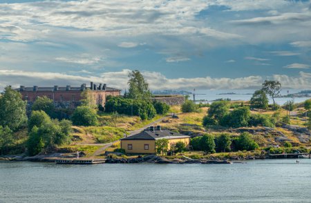 Foto de Helsinki, Finlandia - 20 de julio de 2022: Suomenlinna Fortaleza del mar. Gyllenborg Bastión con murallas y edificio de ladrillo rojo en un entorno verde justo al norte de la Puerta Real. Cloudscape y agua verde claro - Imagen libre de derechos