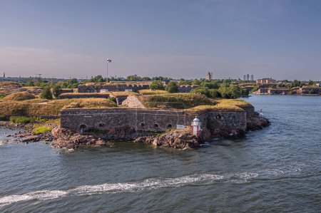 Foto de Helsinki, Finlandia - 20 de julio de 2022: Suomenlinna Fortaleza del mar. Bastión Gustavssvard murallas amplio paisaje verde con faro de luz y murallas bajo el cielo azul y el mar gris - Imagen libre de derechos