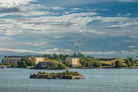 Foto de Helsinki, Finlandia - 20 de julio de 2022: Suomenlinna Fortaleza del mar. Los edificios amarillos y el terreno de la Academia Naval o Pikku Musta isla bajo el paisaje nublado azul. Islote en el mar en frente - Imagen libre de derechos