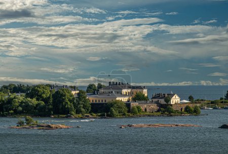 Foto de Helsinki, Finlandia - 20 de julio de 2022: Suomenlinna Fortaleza del mar. Murallas y edificios amarillos y el terreno de la Academia Naval o Pikku Musta isla bajo el paisaje nuboso azul. Isletas en el mar - Imagen libre de derechos