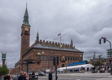 Téléchargez les photos : Copenhague, Danemark - 23 juillet 2022 : Grande mairie de briques rouges avec horloge et clocher domine derrière la place Radhuspladsen très fréquentée sous un paysage nuageux gris. Personnes, voitures et tentes - en image libre de droit