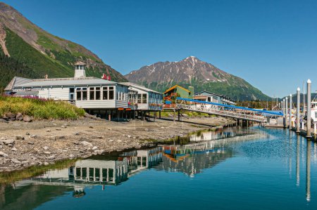 Photo for Seward, Alaska, USA - July 22, 2011: White wooden building of Rays Restaurant and bar along shoreline is mirrored in blue Resurrection Bay water. Blue sky and green mountain flank - Royalty Free Image