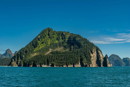 Photo for Resurrection Bay, Alaska, USA - July 22, 2011: Large and tall offshoot forested rock at Thumb Cove bay under blue sky. All green behind azure water with brown rocky cliffs - Royalty Free Image