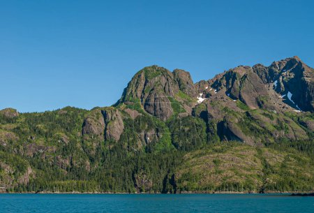 Foto de Resurrection Bay, Alaska, EE.UU. - 22 de julio de 2011: El follaje verde lucha para cubrir el flanco de la costa rocosa rocosa marrón-gris bajo el cielo azul y detrás del agua azul. Pocos parches de nieve - Imagen libre de derechos