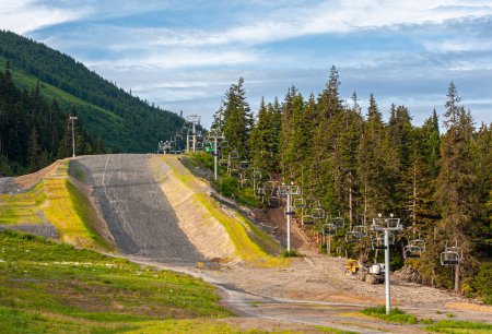 Photo for Girdwood Alaska, USA - July 23, 2011: Ski lift goes up the mountains from Alyeska Resort. Summer shows green trees, forested mountains, gray road, under blue cloudscape. - Royalty Free Image