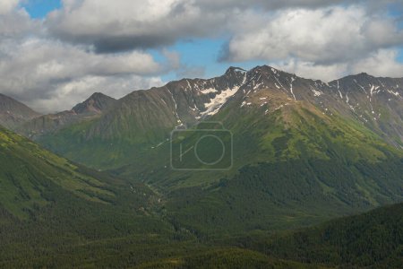 Photo for Girdwood Alaska, USA - July 23, 2011: Chugach Park. Wide mountain range with snow patches and green forested flanks under thick blue cloudscape above town - Royalty Free Image