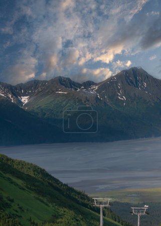 Photo for Girdwood Alaska, USA - July 23, 2011: Blue cloudscape above mountain range with snow patches. 2 ski lift poles up front on green forested flank - Royalty Free Image