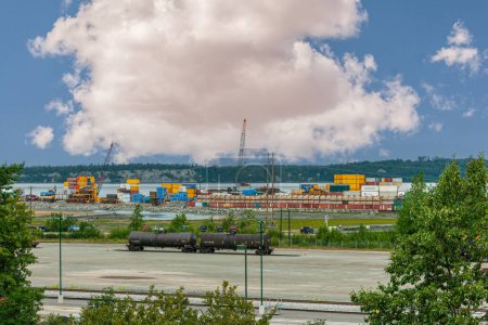 Photo for Anchorage, Alaska, USA - July 23, 2011: 2 black tank train wagons with container stacks in back under thick white cloud set in blue sky. Green foliage and harbor view - Royalty Free Image