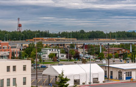 Photo for Anchorage, Alaska, USA - July 23, 2011: Alaska Railroad Corporation red building, short clock tower and other buildings and cars in port area under blue cloudscape. Green foliage sprinkled - Royalty Free Image
