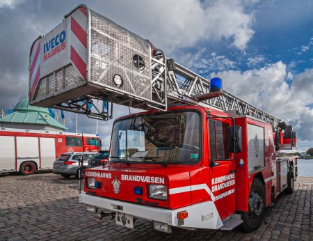 Photo for Copenhagen, Denmark - September 13, 2010: Red and white ladder fire truck parked on harbor quay with other cars under blue cloudscape. Names on truck, IVECO MAGIRUS - Royalty Free Image
