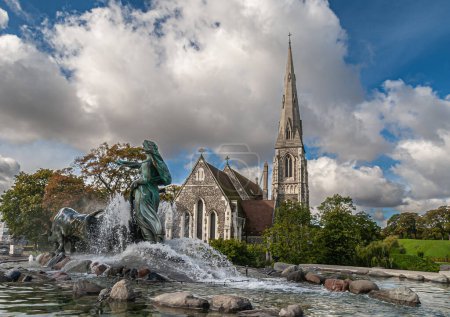 Photo for Copenhagen, Denmark - September 13, 2010: Bronze Gefion Fountain seen from NE over water in front of gray stone Saint Albans church under blue cloudscape landscape - Royalty Free Image