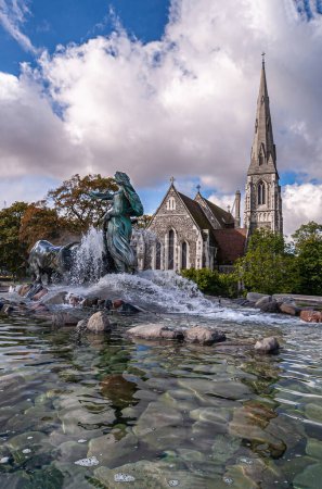 Photo for Copenhagen, Denmark - September 13, 2010: Bronze Gefion Fountain seen from NE over water in front of gray stone Saint Albans church under blue cloudscape - Royalty Free Image