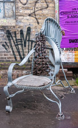 Téléchargez les photos : Copenhague, Danemark - 14 septembre 2010 : Christiania Freetown. Gros plan de chaise de jardin métallique gris artistiquement construit contre mur de briques marron plâtré avec des graffitis et des affiches colorées - en image libre de droit