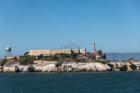 San Francisco, CA, USA - July 13, 2023: West shore of Alcatraz Island showing the main building, water tower and lighthouse. Shoreline covered by white guano under blue sky