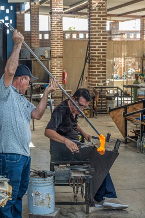 Photo for Mexico, Cabo San Lucas - July 16, 2023: La Fabrica de Vidrio Soplado. Hand blown artistic glass factory. Team of glassblowers melt 2 hot pieces together handling blowpipes and shears - Royalty Free Image