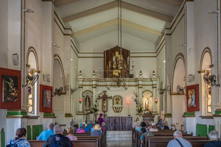 Photo for San Jose del Cabo Centro, Mexico - July 16, 2023: Mission Church. Looking at chancel from back at entrance. People praying on benches. Altar, lanterns and paintings on walls - Royalty Free Image