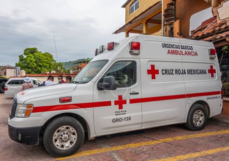 Foto de Zihuatanejo, México - 18 de julio de 2023: Furgoneta ambulanc rojo-blanca estacionada al final del muelle bajo un paisaje nublado gris. Fachada de edificio amarillo como telón de fondo. Personas y follaje verde - Imagen libre de derechos