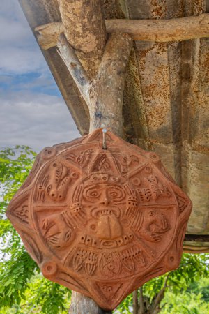 Téléchargez les photos : Zihuatanejo, Mexique - 18 juillet 2023 : Gros plan, œuvre d'art du symbole solaire aztèque relligieux produite dans un four historique en terre cuite accroché le long du tronc d'arbre. Feuillage vert, toit brun, paysage nuageux bleu - en image libre de droit