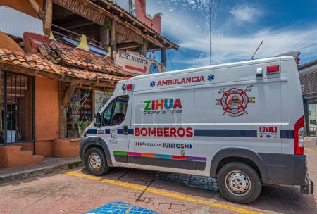 Foto de Zihuatanejo, México - 18 de julio de 2023: Furgoneta de bomberos ambulancia estacionada en el centro de la ciudad frente a un restaurante con paredes rojas y paneles de techo. Diferentes colores sobre fondo blanco. Paisaje nuboso azul - Imagen libre de derechos