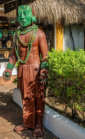 Foto de Guatemala, Puerto Quetzal - 20 de julio de 2023: Primeros planos, estatua masculina de piedra marrón con máscara de dios de jade verde y otras joyas de jade en brazos, dedos y cuello, dando la bienvenida a los turistas en la terminal de cruceros - Imagen libre de derechos