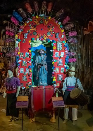 Foto de Guatemala, La Antigua - 20 de julio de 2023: Finca La Azotea. 3 muñecas ambientadas en un paisaje cristiano con un montón de imágenes de Jesús y santos contra la pared roja decorada con plumas y más - Imagen libre de derechos