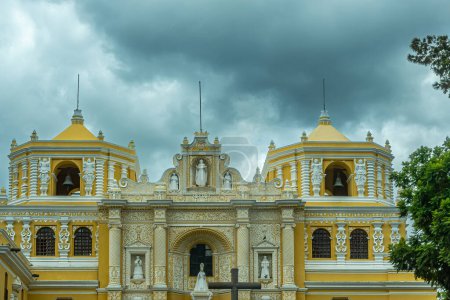 Foto de Guatemala, La Antigua - 20 de julio de 2023: Convento-Iglesia Nuestra Señora de la Merced. Nivel superior con campanarios amarillos y fachada, estatuas blancas en nichos bajo un oscuro paisaje nublado - Imagen libre de derechos