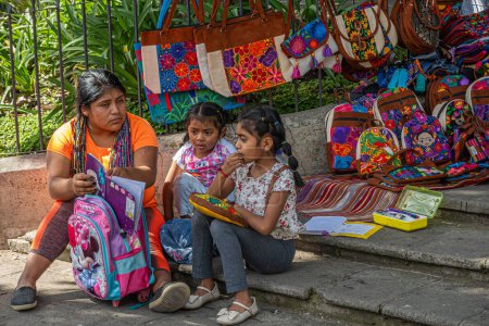 Photo for Guatemala, La Antigua - July 20, 2023: Mother in modern clothing, merchant selling coloful bags and backpacks sits with 2 daughters on step helping her reciting homework. Plaza Mayor, central square - Royalty Free Image