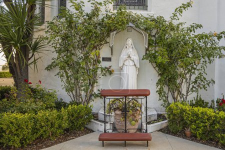 Foto de Santa Barbara, CA, EE.UU. - 19 de enero de 2024: Santa Frances Xavier Cabrini estatua blanca fuera de la iglesia Nuestra Señora de los Dolores, alias Francesca Saverio. Está rodeada de follaje verde. Banco de oración en frente - Imagen libre de derechos