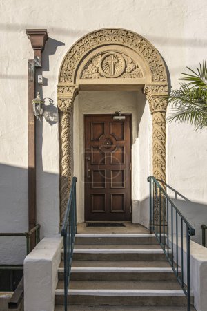 Foto de Santa Barbara, CA, EE.UU. - 19 de enero de 2024: Arco esculpido alrededor de la puerta de entrada trasera marrón reservada para sacerdotes a la iglesia Nuestra Señora de los Dolores. Ángeles y cruz. Set en pared beige pálido - Imagen libre de derechos