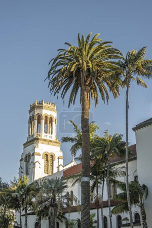 Foto de Santa Bárbara, CA, EE.UU. - 19 de enero de 2024: Torre de Nuestra Señora de los Dolores iglesia iluminada contra el cielo azul. Follaje de palmera verde frente a la fachada oeste - Imagen libre de derechos